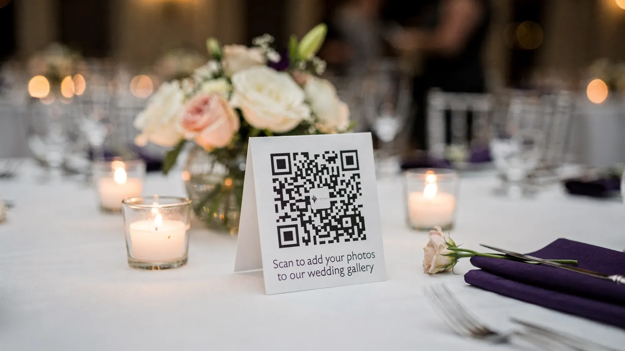 A wedding reception table setting with a small, elegant tent card sign featuring a prominent QR code and a short instruction like “Scan to add your photos to our wedding gallery,” surrounded by candles and simple floral decor.