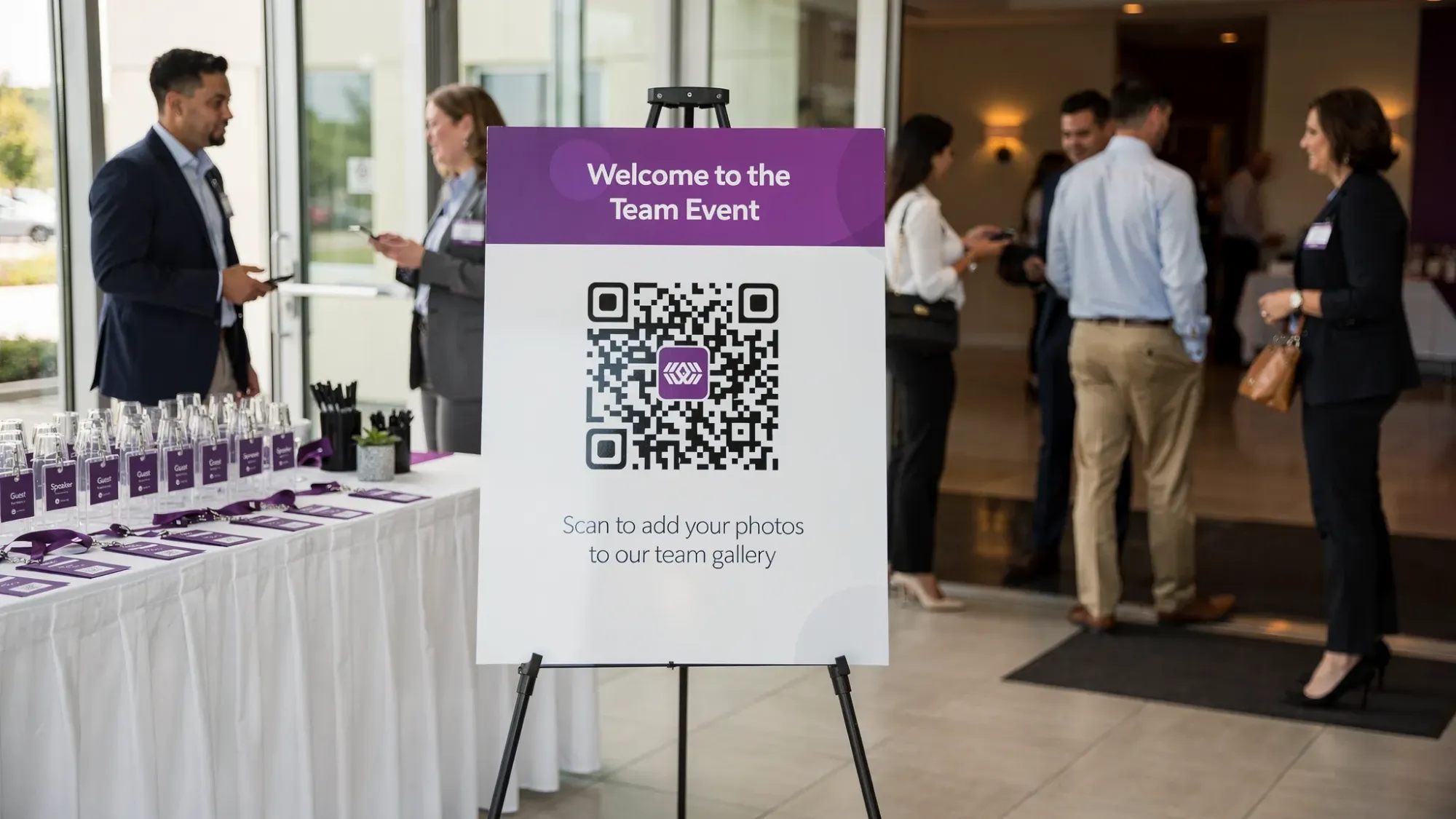 A corporate event photo collection setup at a venue entrance: a clean welcome sign on an easel with a large QR code and short copy inviting guests to scan and add photos to the team gallery. Nearby is a registration table with badges and lanyards, and people arriving in business casual attire.