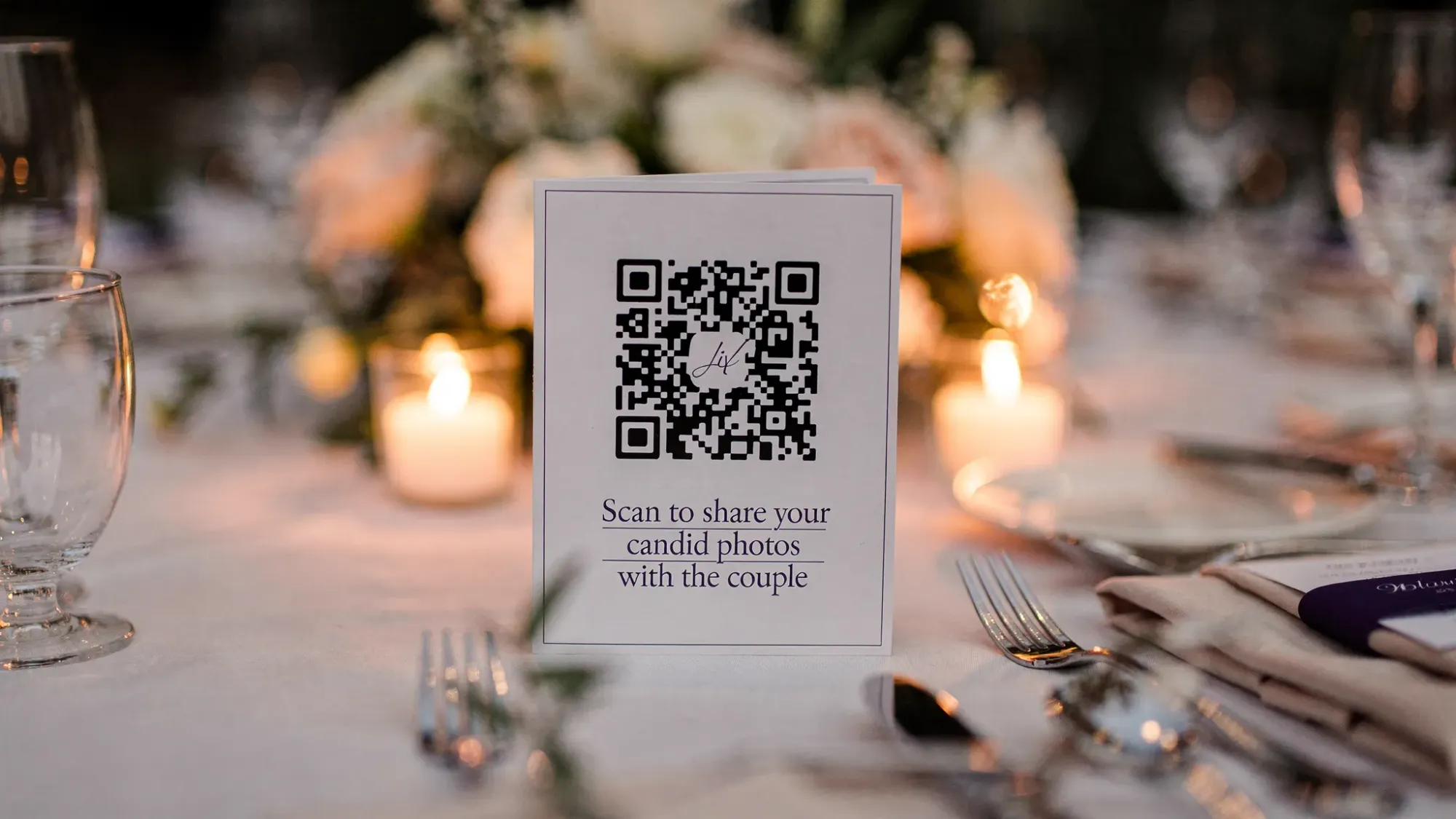 A wedding reception table with a simple table tent featuring a QR code and short text inviting guests to scan to take candid photos for the couple, with candles and floral decor in the background.