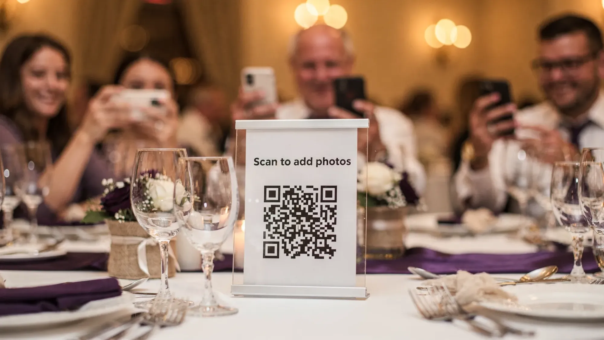 A wedding reception table with a simple sign that says “Scan to add photos” next to a QR code, while guests nearby use their phones to scan and take candid photos. The scene includes warm reception lighting, glassware, and a celebratory atmosphere.