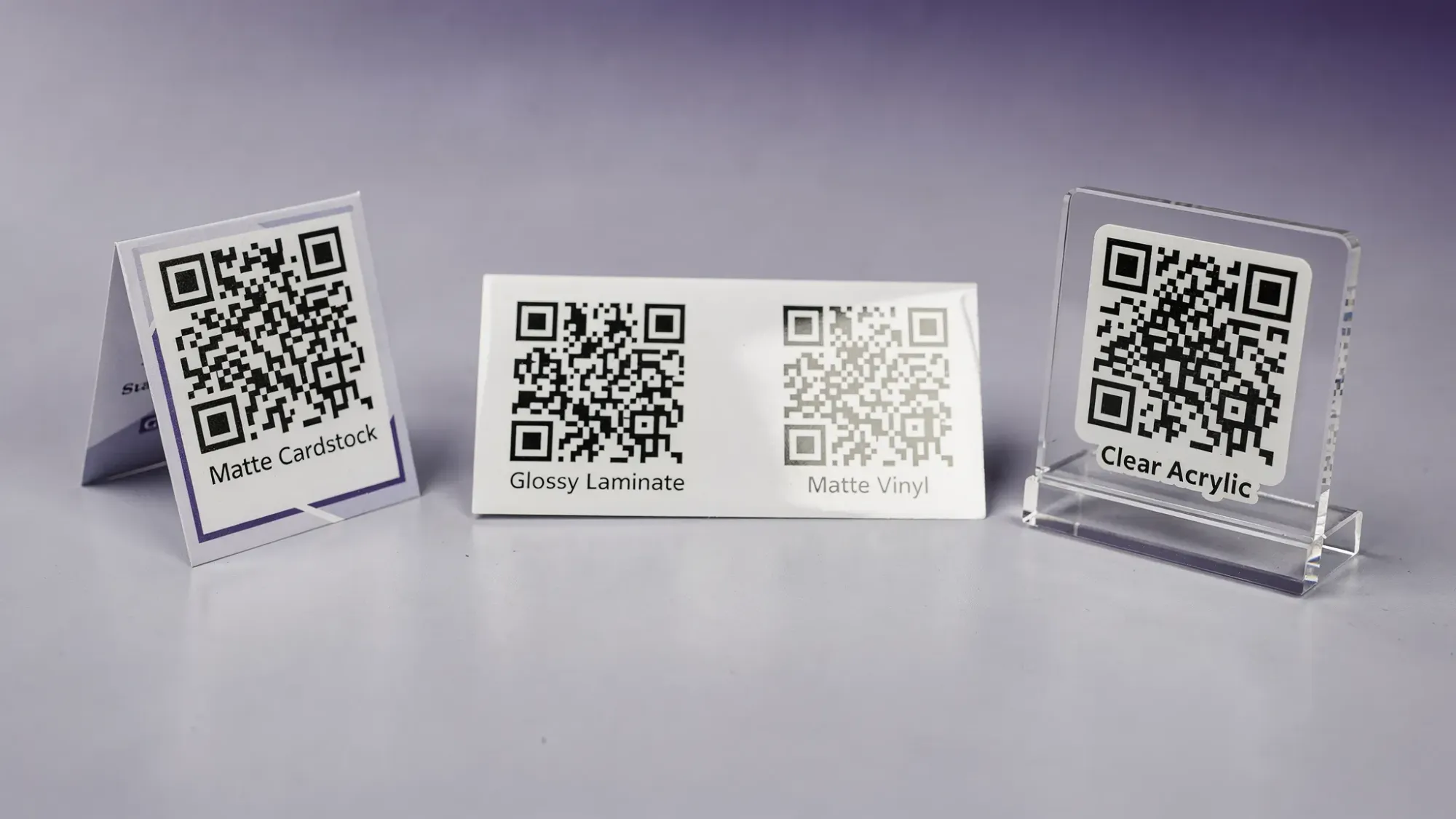 A side-by-side close-up of common event QR tag materials (matte cardstock table tent, glossy laminated card, matte vinyl sticker, clear acrylic sign) under overhead light, showing how glare affects scan readability.