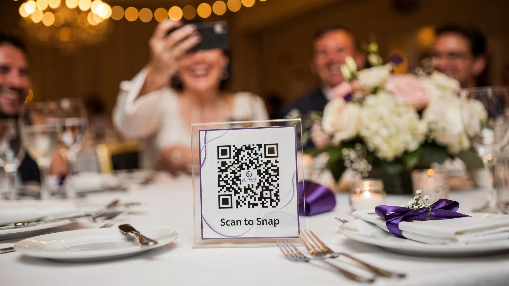 A wedding reception table with a small acrylic table tent displaying a QR code that says “Scan to Snap,” guests laughing in the background while one person holds up a phone to scan, warm ambient lighting and floral centerpiece visible.