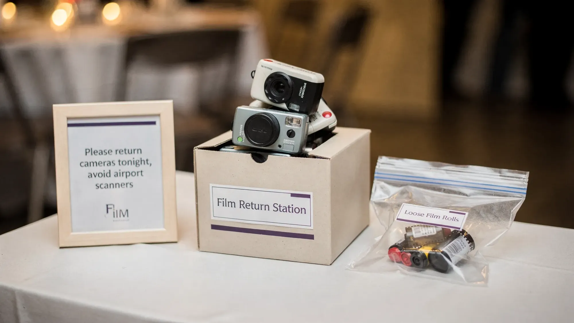 A small event “film return” station with a labeled collection box for disposable cameras, a simple sign that says ‘Please return cameras tonight, avoid airport scanners’, and a sealed zip pouch for any loose rolls.
