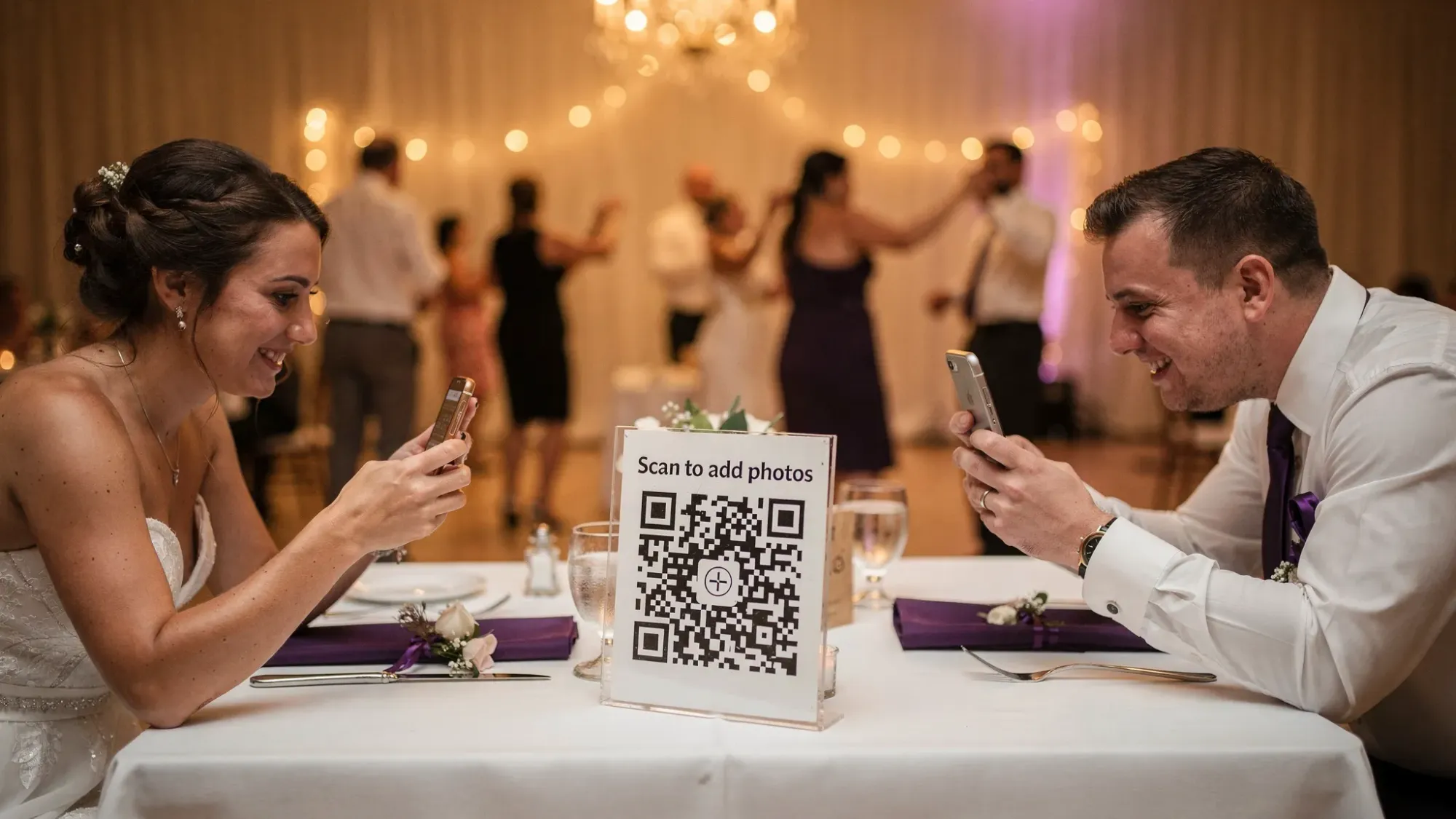 A wedding reception table with a simple, elegant QR-code sign that says “Scan to add photos,” while two guests hold phones up and scan the code; in the background, a lively dance floor and warm reception lighting.