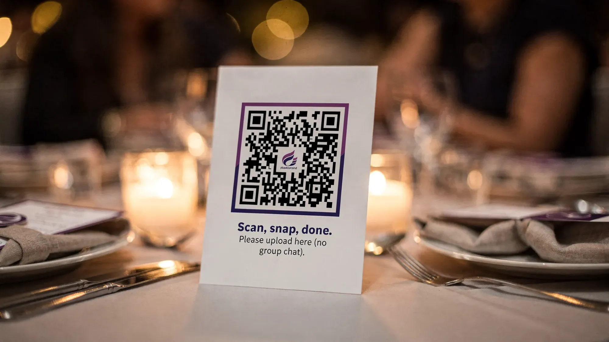 A wedding reception table tent next to place settings and candles, featuring a large QR code and short copy that says “Scan, snap, done. Please upload here (no group chat).” The scene shows warm reception lighting and a clean, modern sign design.