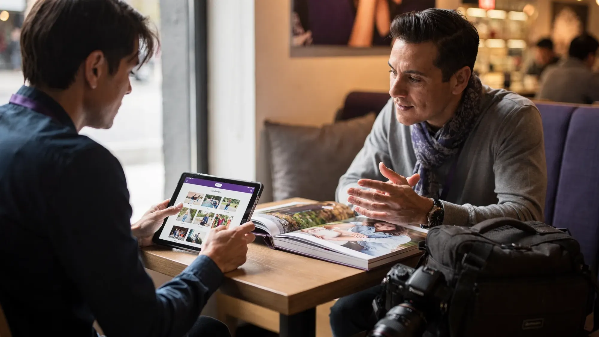 A client reviewing a photographer’s portfolio on a tablet during a consultation at a cafe table. The photographer has a camera bag nearby and a printed sample album open, creating a professional planning vibe.