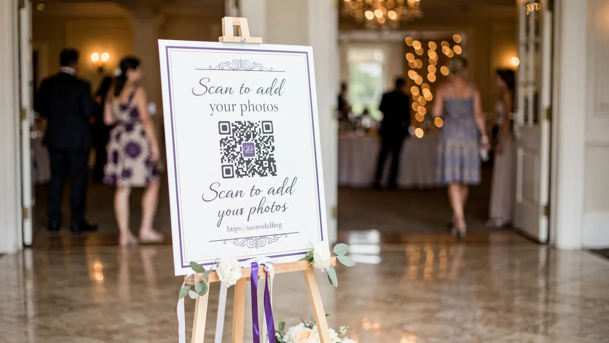 A wedding reception welcome sign on an easel with a large QR code and short URL that says “Scan to add your photos,” placed near the entrance with guests walking by in the background.