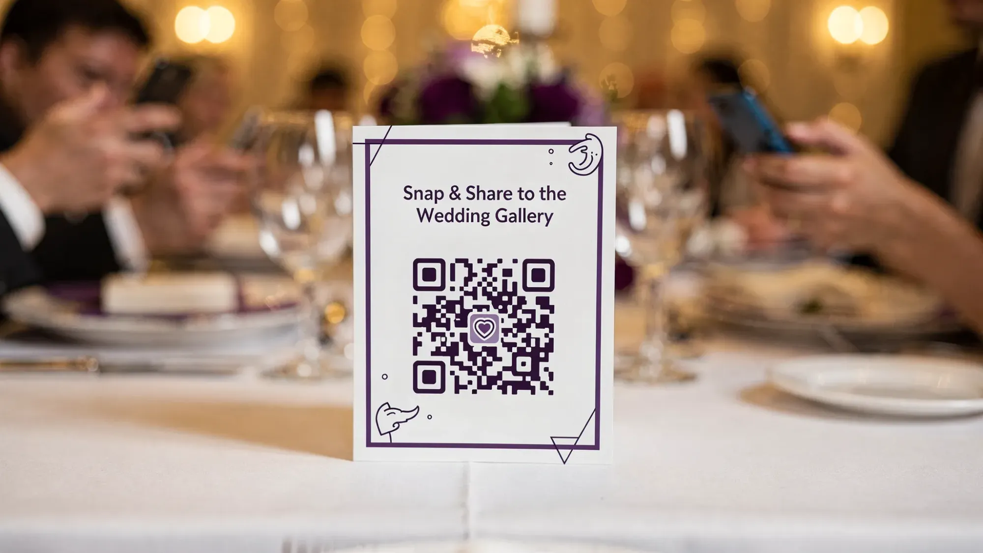 A wedding reception table tent displaying a QR code labeled “Snap & Share to the Wedding Gallery,” with guests holding phones nearby in a softly lit venue.