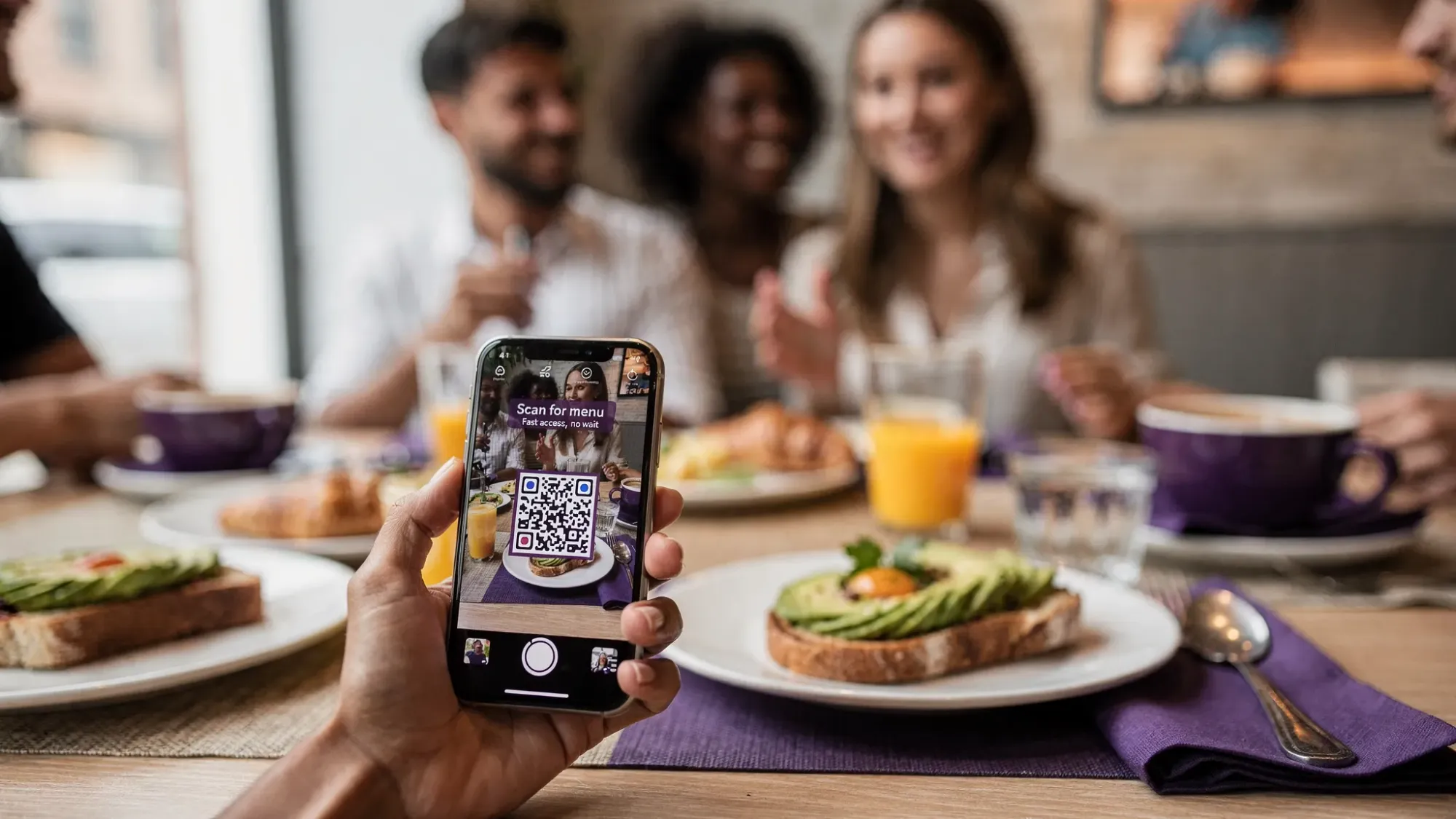 A close-up of a guest scanning a printed QR code with a smartphone camera at a brunch table, with friends in the background and the phone screen facing the camera direction, emphasizing quick access and real-world use.