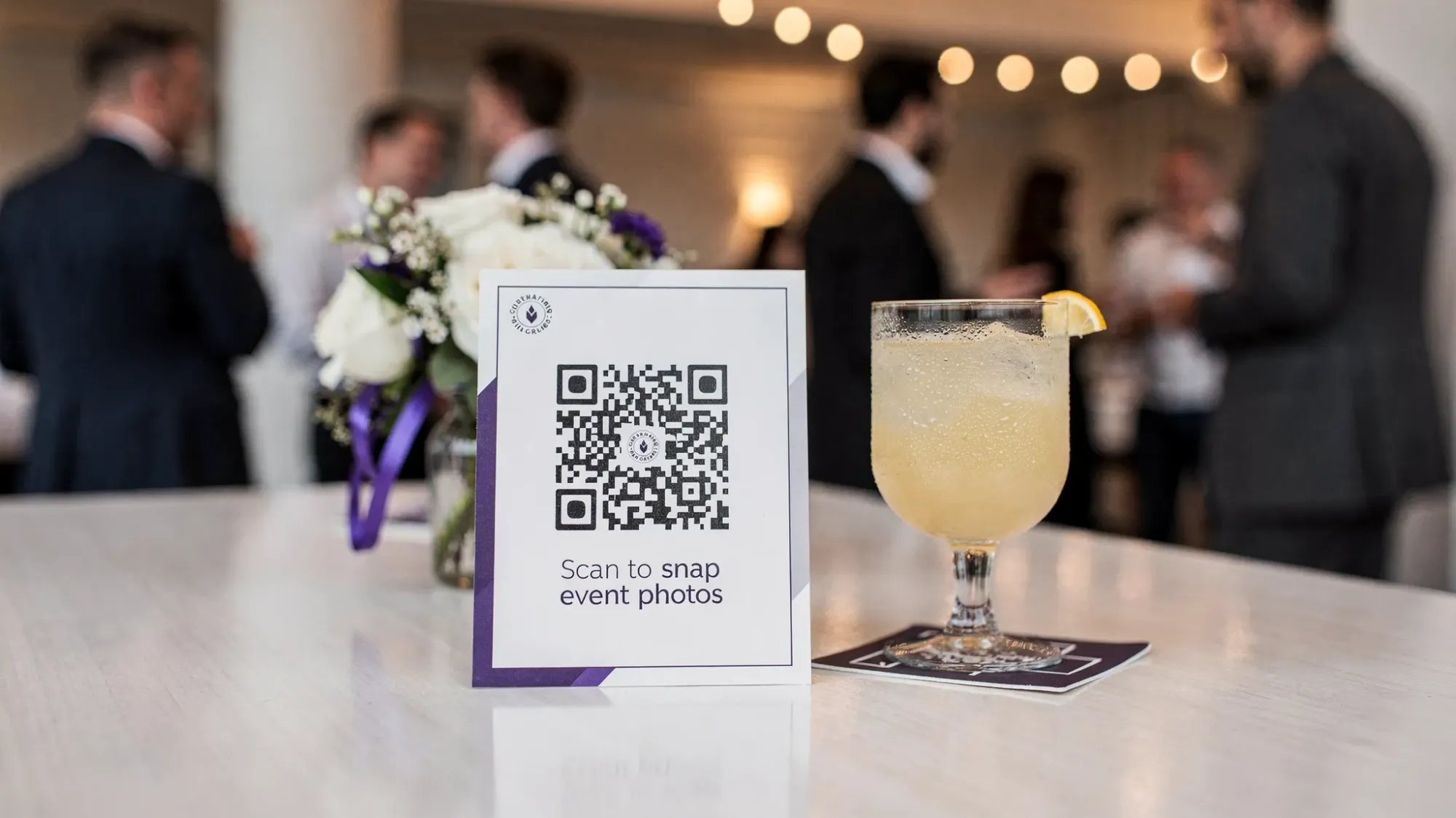 A small table tent sign at an event with a prominent QR code and simple text “Scan to snap event photos,” placed next to a drink and a centerpiece, with guests mingling softly in the background.