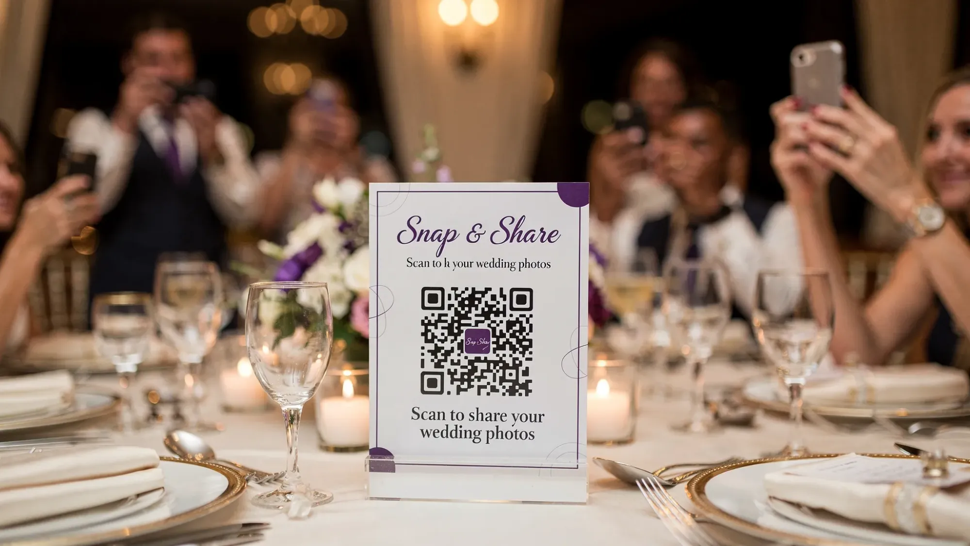 A wedding reception table with a small QR code sign that says “Snap & Share” and guests holding phones taking candid photos, with the QR code facing the camera and readable.