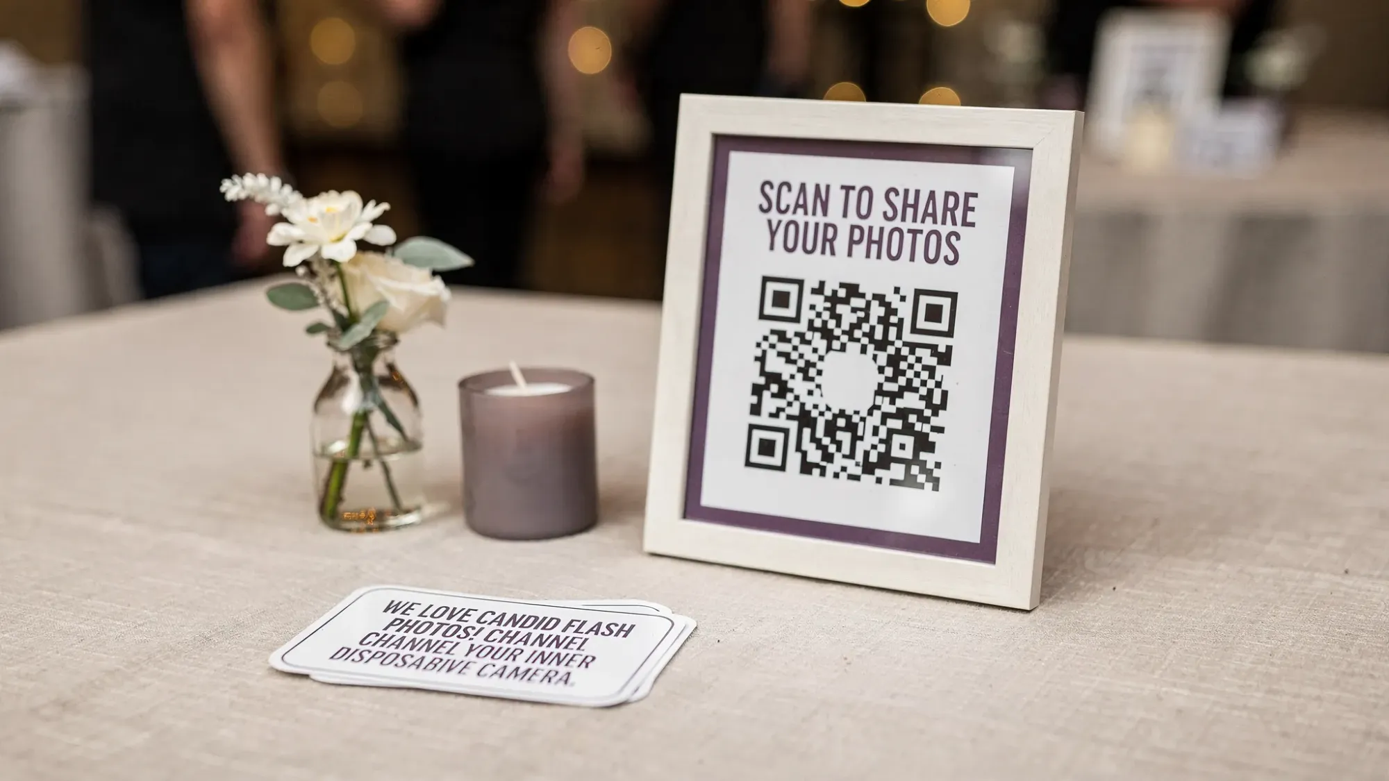 A tabletop event setup with a small sign showing a QR code for guests to scan, placed next to minimalist decor and a note encouraging candid flash photos, evoking a disposable camera theme.