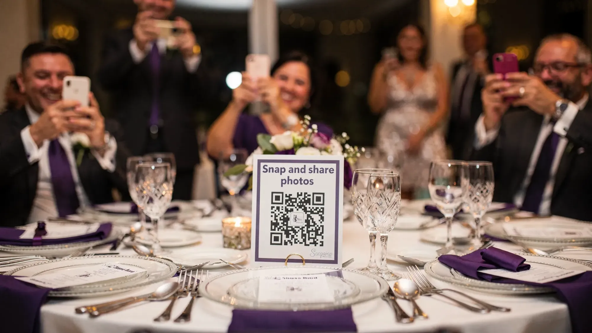 A wedding reception table with a small table tent displaying a QR code labeled “Snap and share photos,” with guests in the background holding phones and taking candid pictures.
