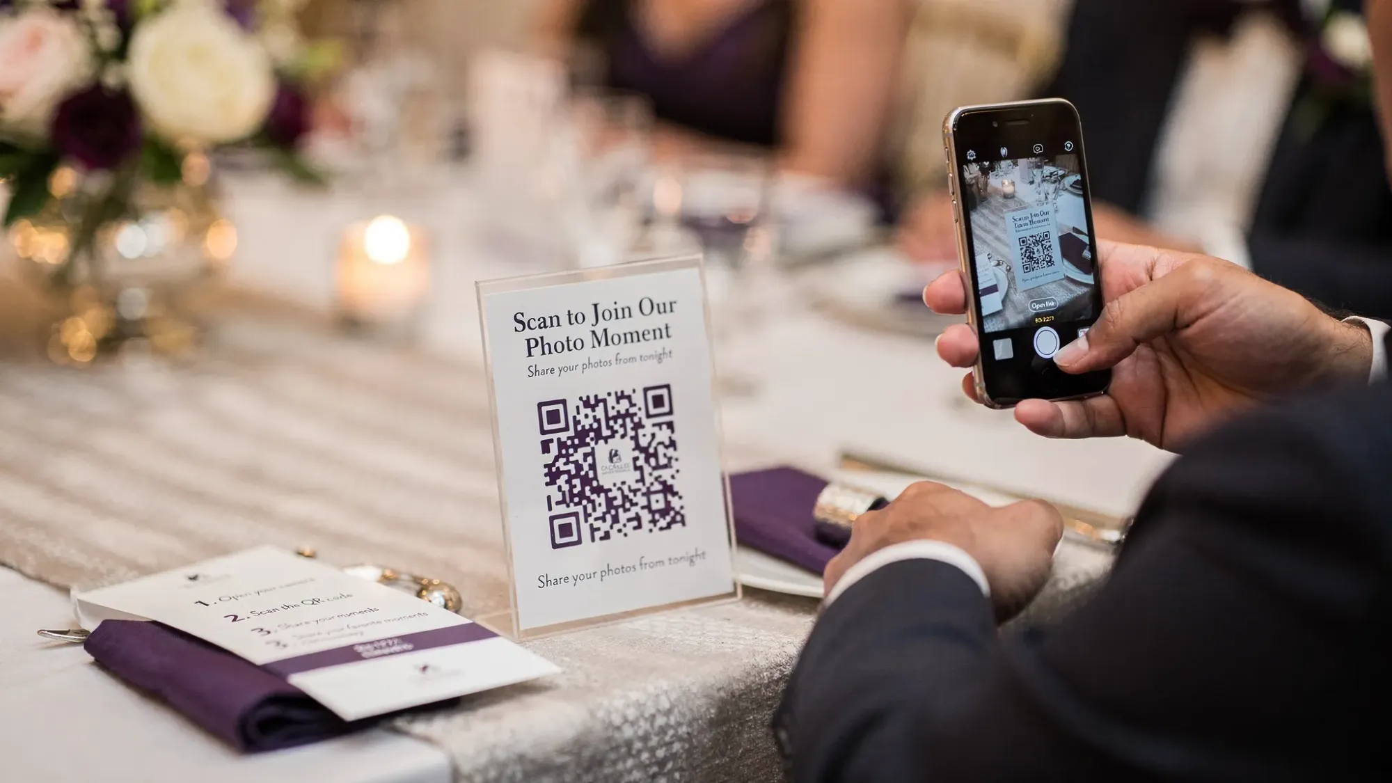A printed QR code sign on a wedding reception table next to a simple instruction card. A guest holds a phone with the camera app open, scanning the QR code to join a shared event photo Moment.