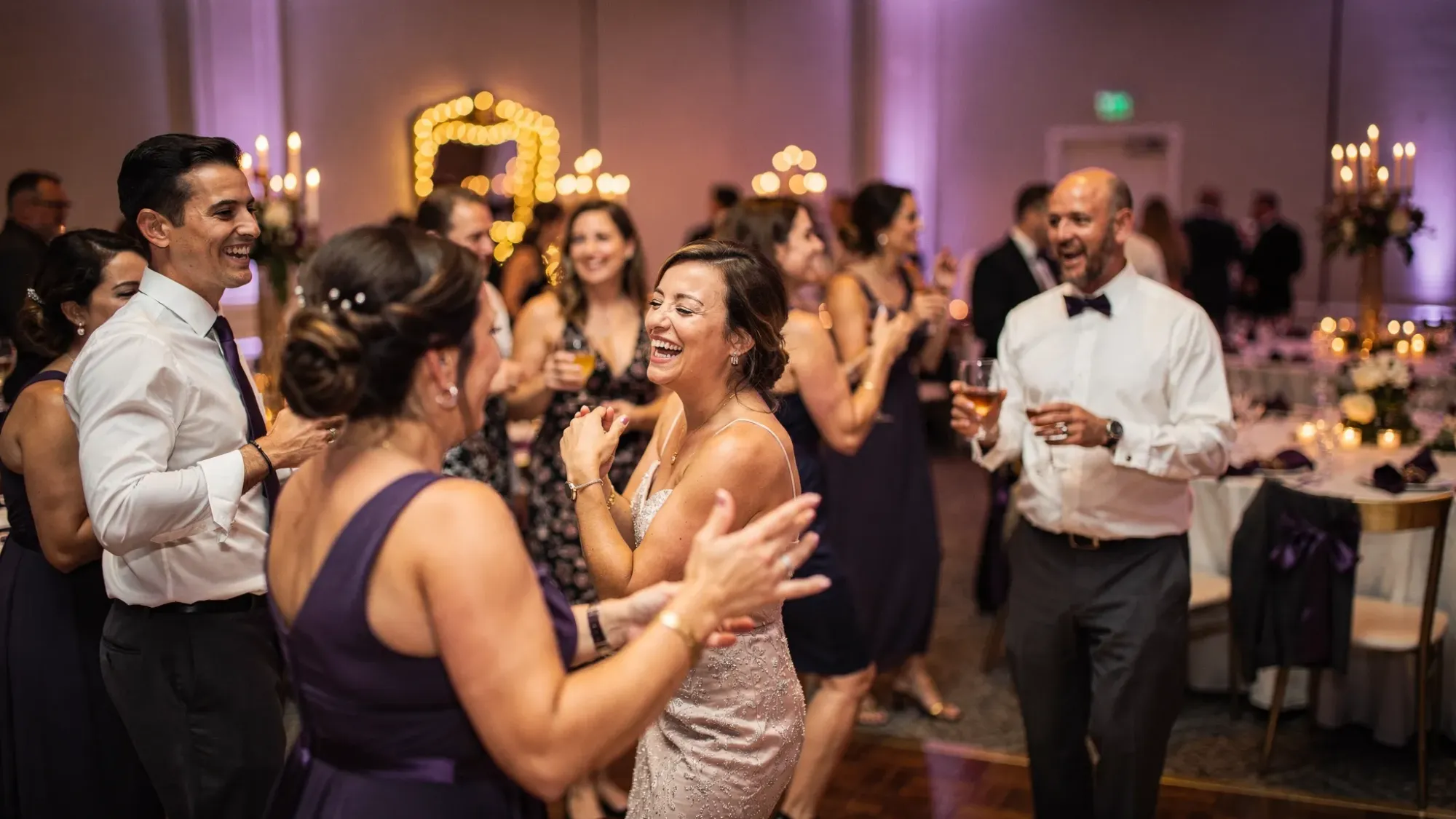 A lively wedding dance floor scene with guests laughing and dancing in warm reception lighting, captured as an authentic candid moment, with no one posing for the camera.