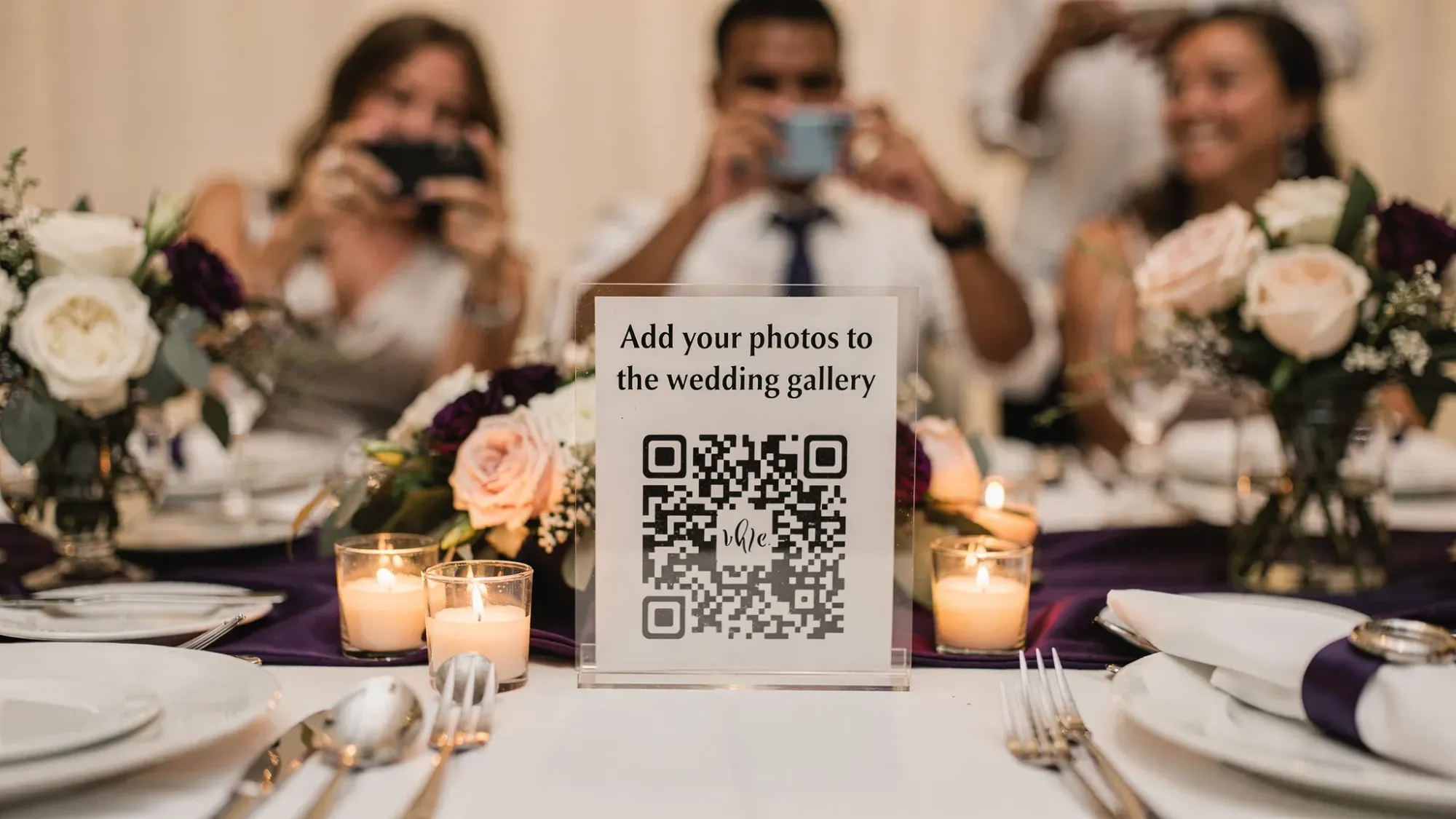 A wedding reception table with a small sign featuring a QR code labeled “Add your photos to the wedding gallery,” surrounded by candles and florals, with guests in the background taking candid photos.