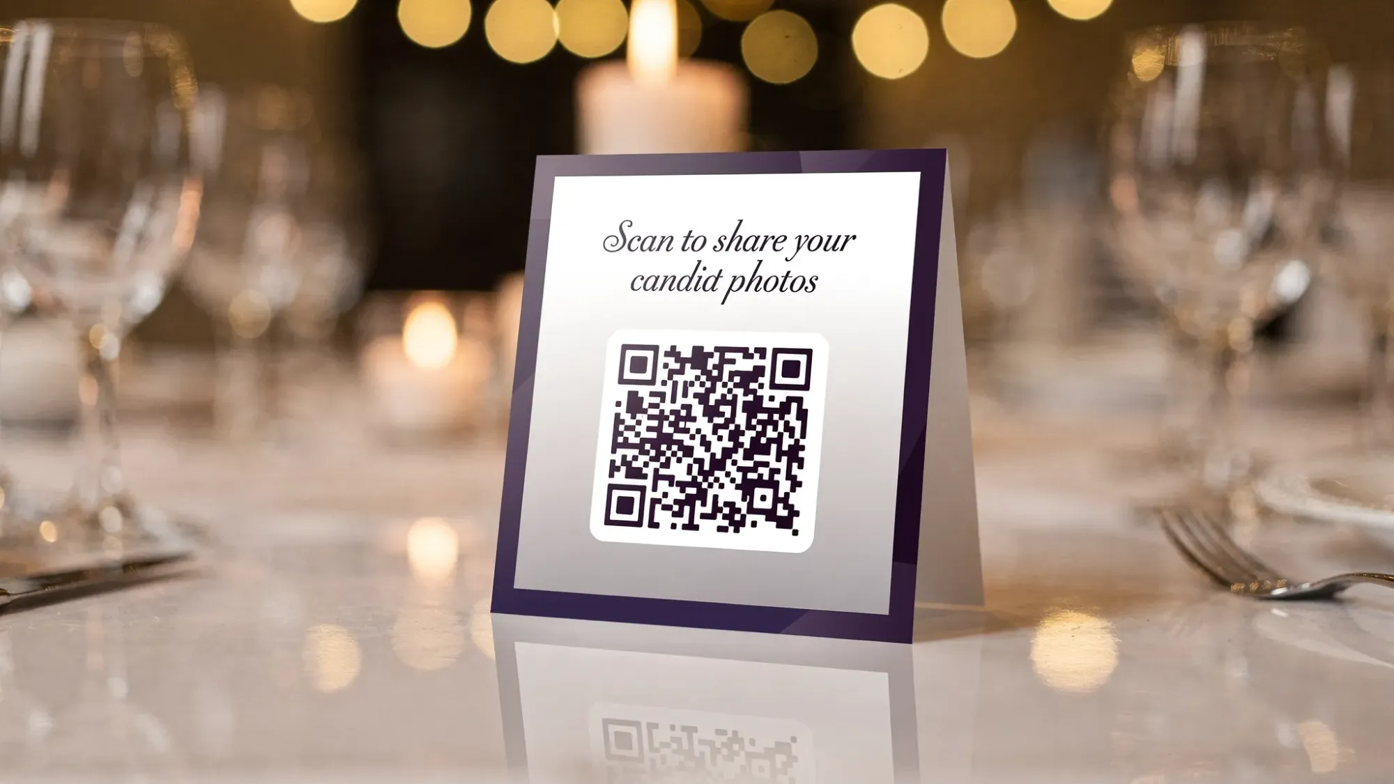 A wedding reception table with a small elegant table tent displaying a QR code and a short line of copy inviting guests to take candid photos, with warm ambient lighting and glassware in the background.
