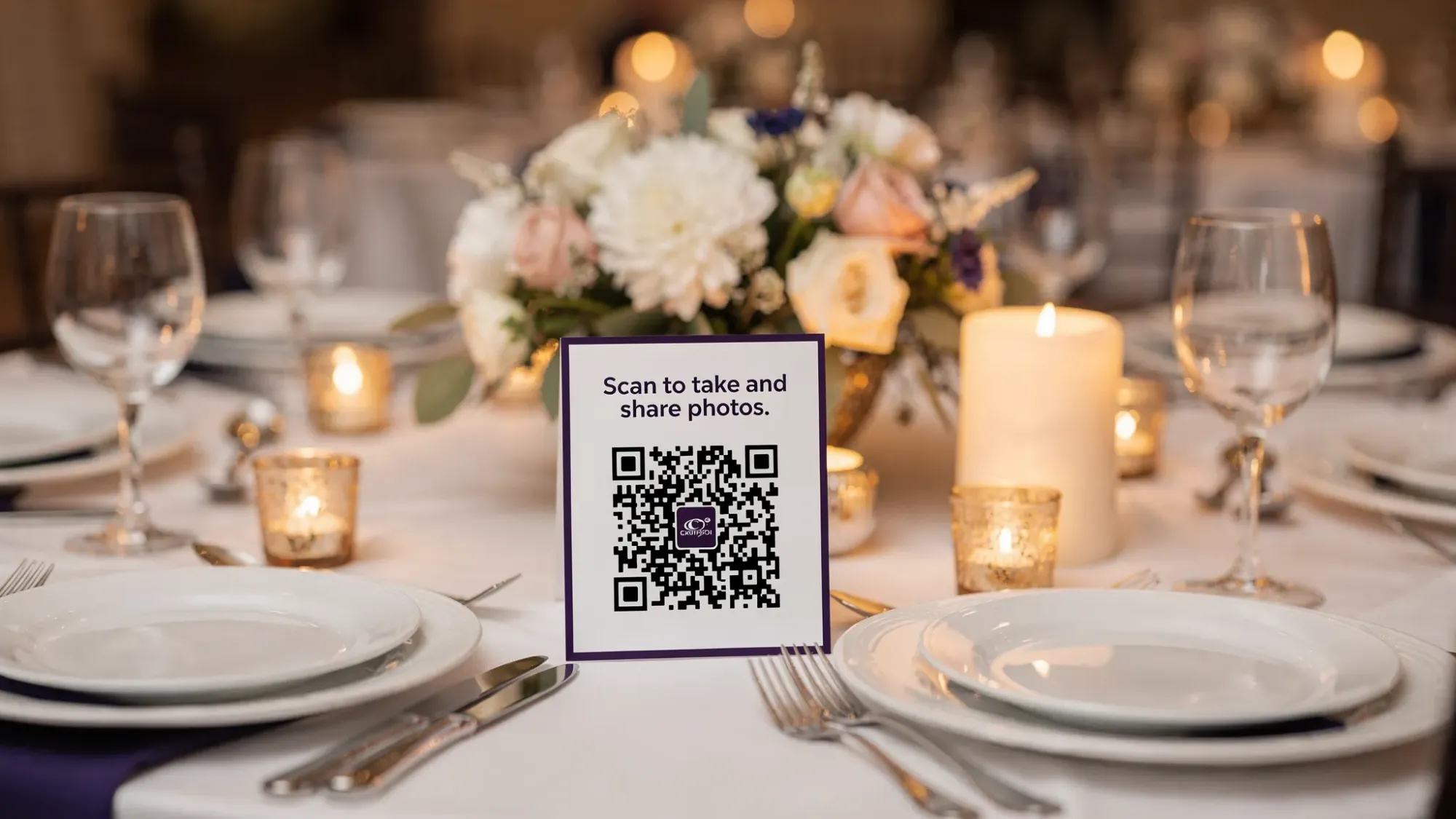 A wedding reception table with a small, elegant table tent displaying a QR code and short text inviting guests to “Scan to take and share photos.” Nearby are place settings, soft candlelight, and a floral centerpiece, suggesting a real event environment.