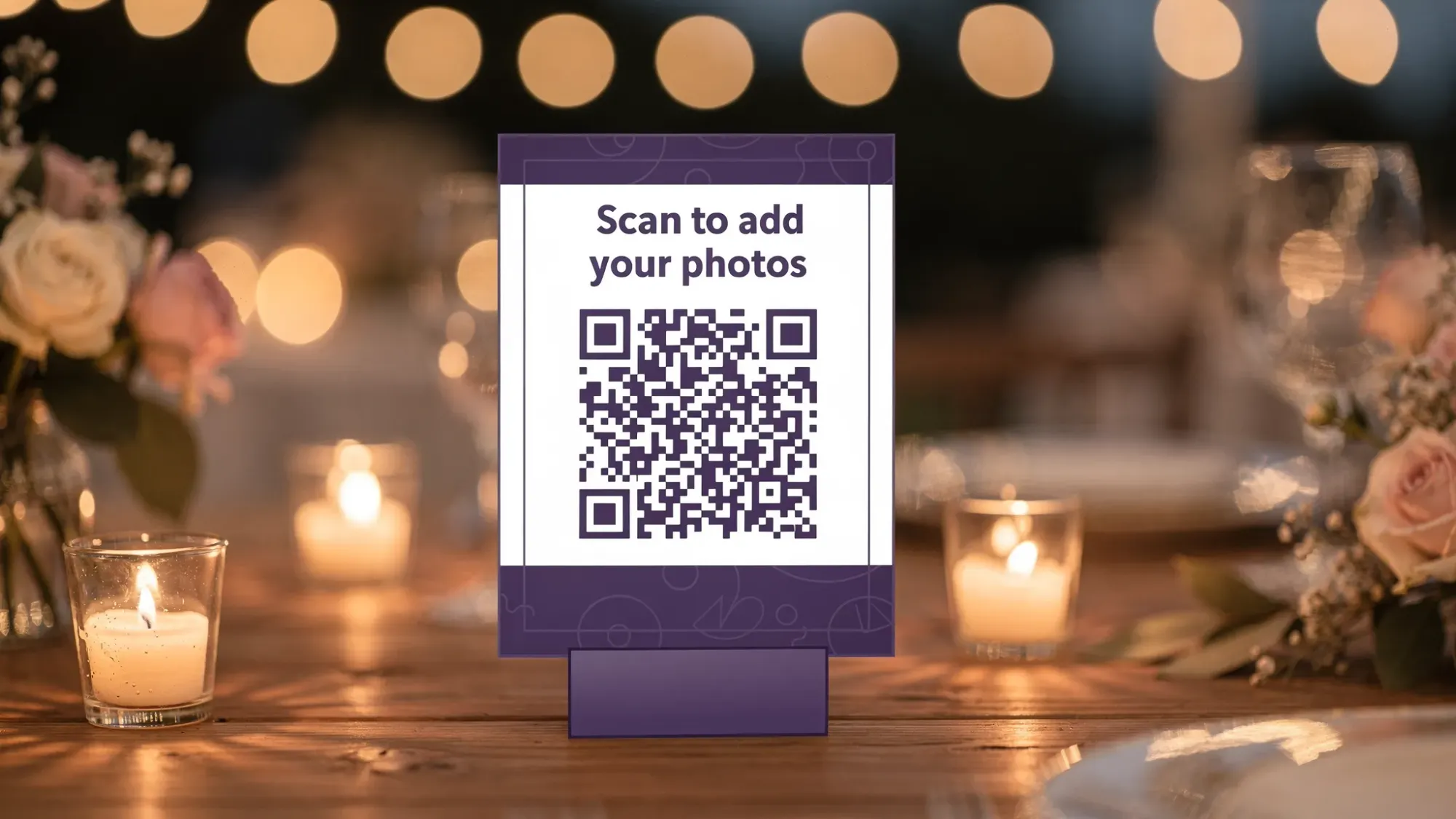 A small wedding table sign with a bold QR code and simple instructions to “Scan to add your photos,” placed next to candles and reception decor in warm evening light.