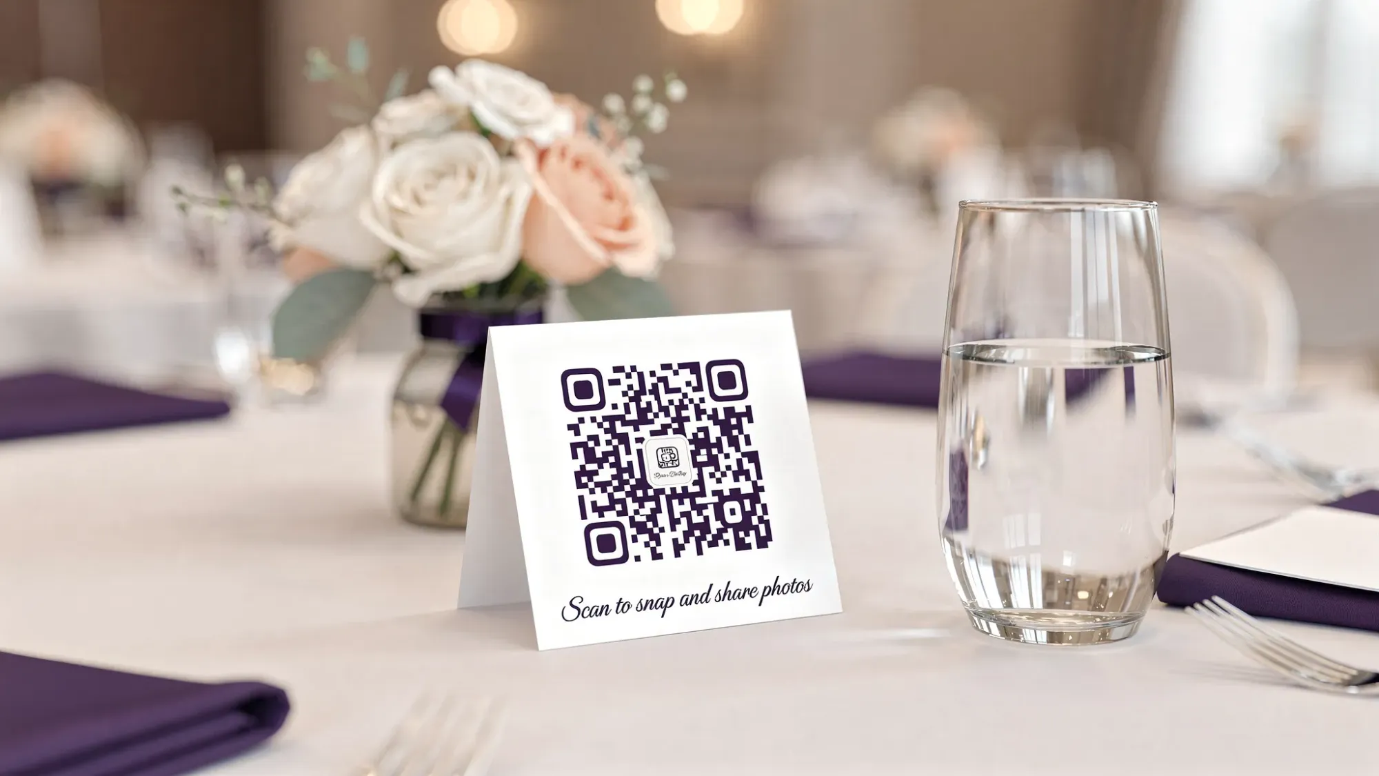 A wedding reception table setting with a small tent card displaying a QR code and short text inviting guests to “Scan to snap and share photos,” placed beside a centerpiece and water glass.