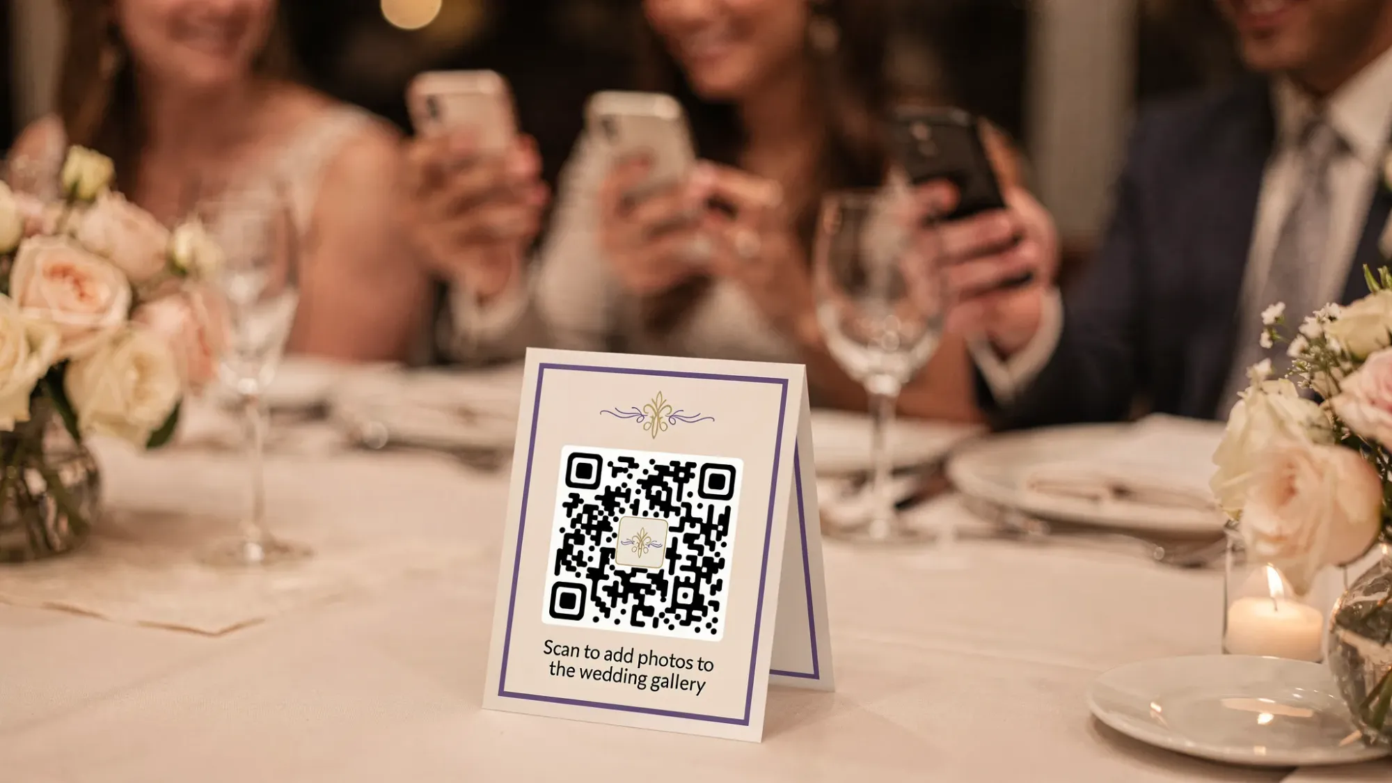 A wedding reception table with a small table tent displaying a QR code and a short instruction like “Scan to add photos to the wedding gallery,” with guests holding phones nearby in a natural candid moment.