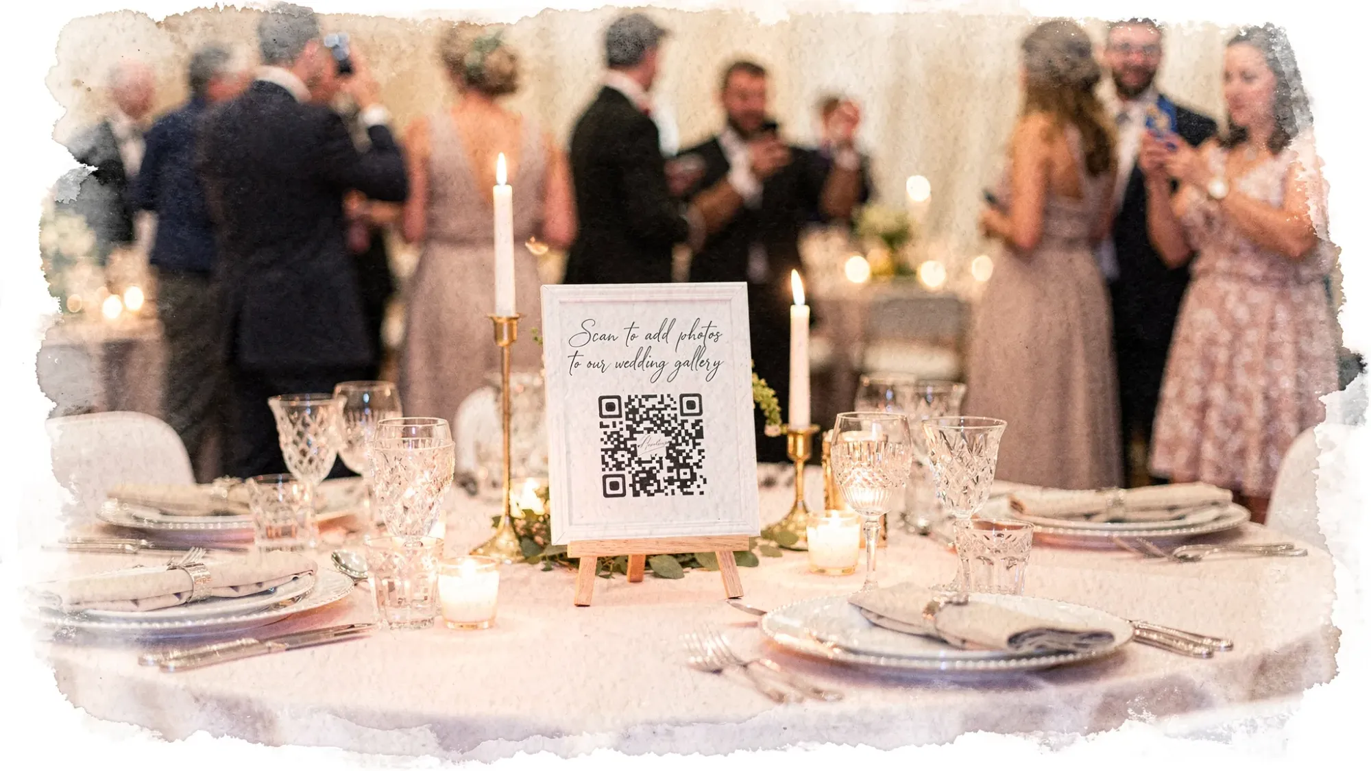 A wedding reception table with a simple framed sign that says “Scan to add photos to our wedding gallery” and a large QR code. Nearby are place settings, candles, and soft ambient lighting. Guests in the background are mingling and taking candid photos.