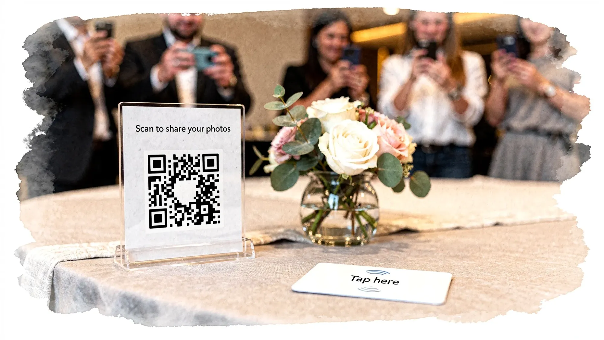 A stylish event table with a small QR code sign, an NFC tap card, flowers, and guests in the background taking candid photos on their phones.