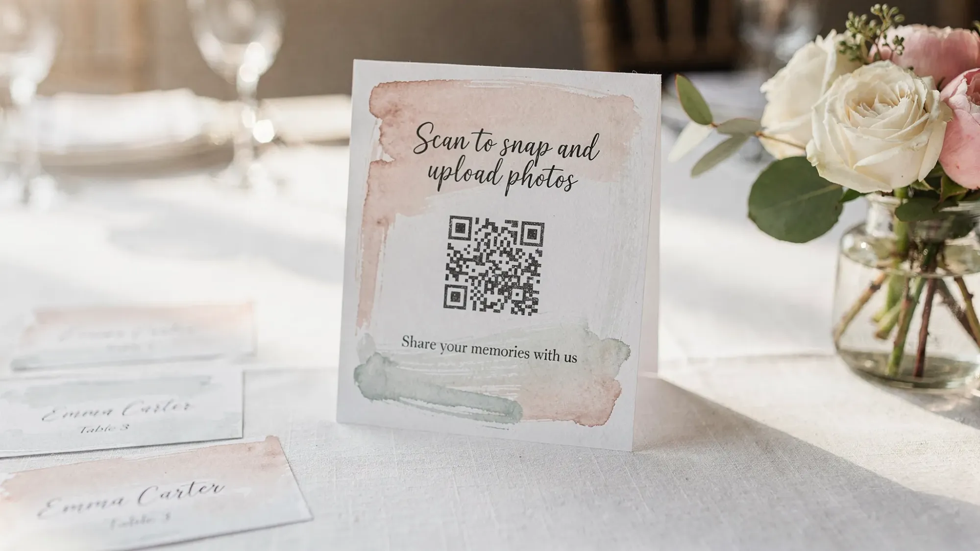A wedding table tent sign with a QR code inviting guests to “Scan to snap and upload photos,” placed next to place cards and a small floral centerpiece.