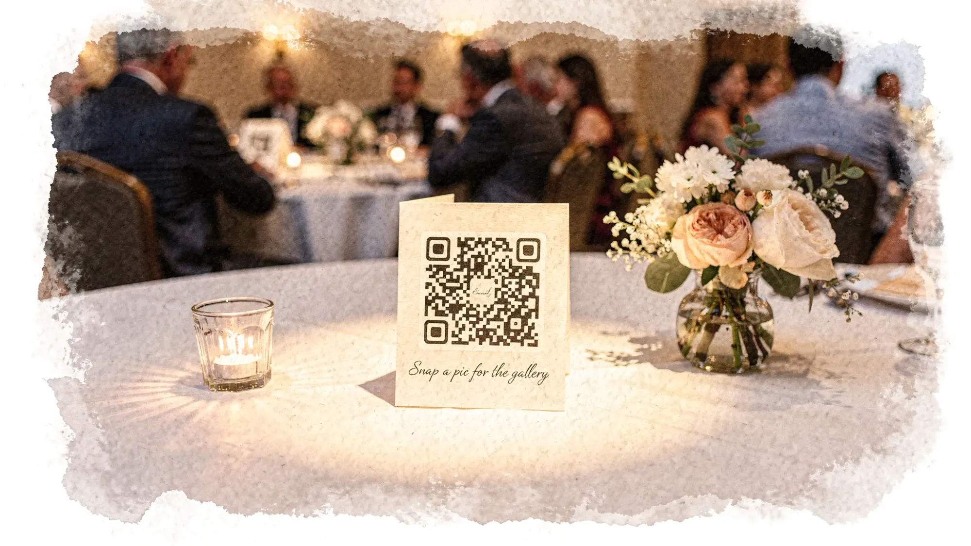 A wedding reception table tent featuring a QR code labeled “Snap a pic for the gallery,” placed near a candle and small centerpiece in a dimly lit venue. Guests are visible in the background, and the QR sign is positioned in a well-lit spot to encourage participation and easy scanning.